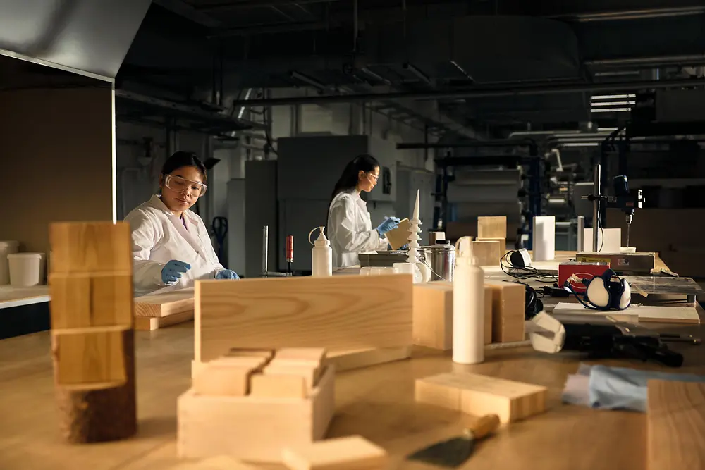 Workshop setting with individuals wearing protective lab coats and gloves, working at a large wooden table covered with wood samples, tools, and bottles.
